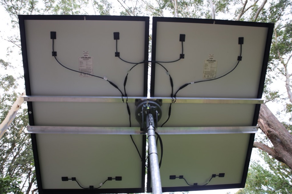 The rear of a solar array mounted on a tilting frame in a garden, showing its wiring and pole mount